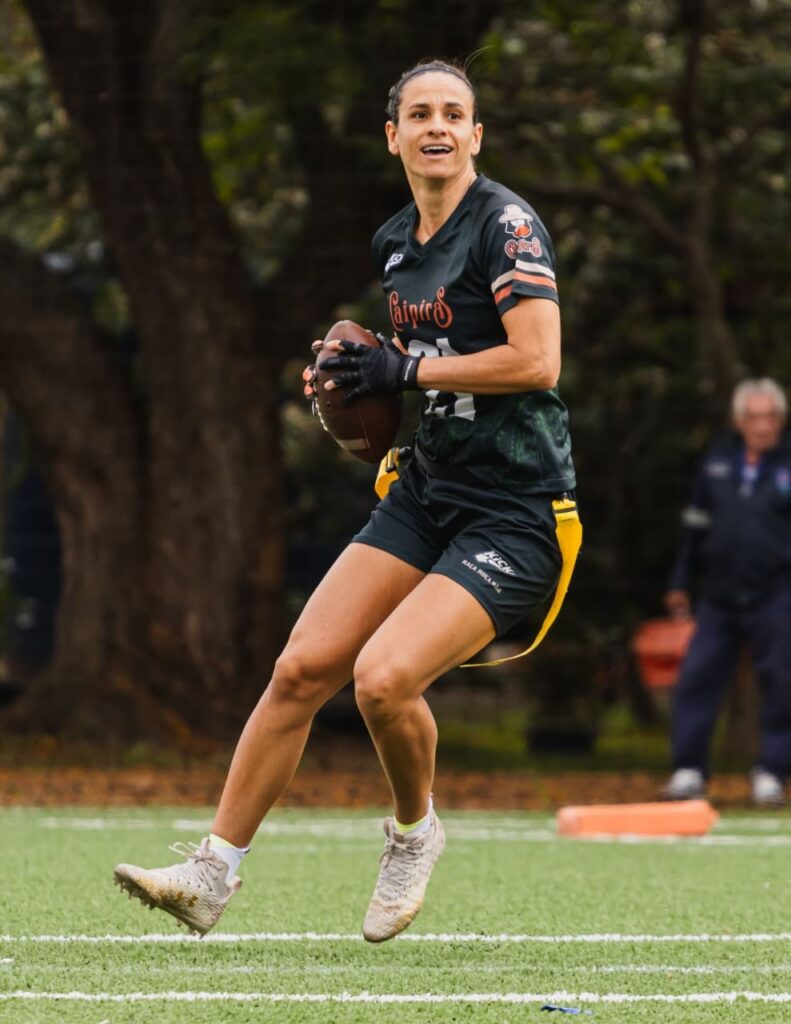 A imagem mostra uma jogadora de futebol americano em ação, segurando uma bola oval e com uma expressão focada no rosto. Ela está usando um uniforme escuro com detalhes em laranja e branco nas mangas, shorts pretos com uma faixa amarela e tênis de futebol. As pernas da jogadora estão em movimento, indicando que ela está correndo ou se preparando para um passe. O campo é gramado e verde, com linhas brancas demarcando o jogo. Ao fundo, pode-se ver árvores e uma pessoa observando o jogo.