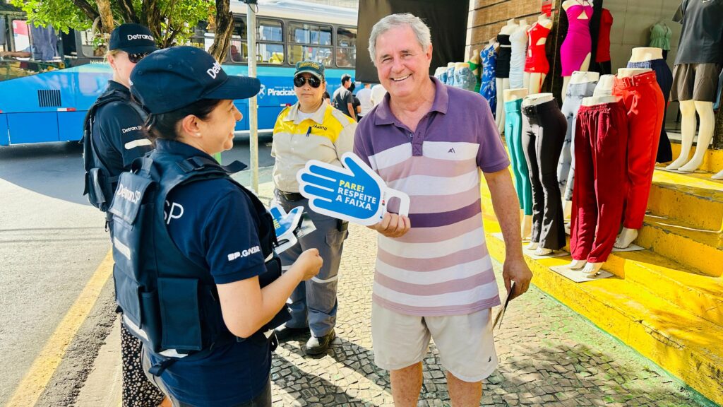 A imagem mostra um homem sorridente em primeiro plano, segurando um sinal em forma de mão azul com a inscrição "PARE! RESPEITE A FAIXA." em branco. Ele está vestindo uma camisa polo listrada em tons de roxo e rosa, e shorts brancos. Ao seu lado, há duas mulheres vestindo uniformes escuros com coletes azuis e bonés pretos com o logo "Detran.SP". Uma delas, à esquerda, está falando com o homem e também segura um sinal semelhante. A outra mulher, mais atrás, está usando um uniforme amarelo e cinza com um boné de trânsito. No fundo, vê-se um ônibus azul com a inscrição "Piracicaba". À direita do homem, há uma vitrine de loja com manequins exibindo roupas coloridas, como vestidos e calças, dispostas em degraus amarelos. A cena parece ocorrer em uma rua ensolarada, com o chão de paralelepípedos visível.