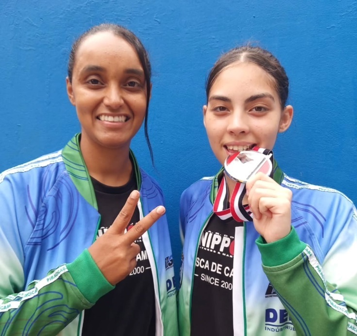 A imagem mostra duas jovens sorrindo para a câmera, ambas usando jaquetas esportivas azuis e verdes. A jovem da esquerda está fazendo o sinal de paz com os dedos e a da direita está segurando uma medalha perto da boca. A medalha tem uma fita listrada em vermelho, preto e branco. Ao fundo, uma parede azul. As jaquetas têm logotipos e texto que parecem indicar uma equipe ou evento esportivo, com menções a "PIRACICABA" e "DESDE 2000".