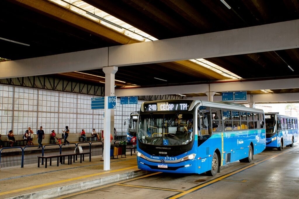 A imagem mostra um terminal de ônibus com dois ônibus azuis estacionados em plataformas. O ônibus da frente, com o número 240 e destino "CECAP / TPI", está mais próximo do espectador e é o foco principal. Ele é um ônibus moderno da marca Iveco, com janelas amplas e um design aerodinâmico. No fundo, pessoas estão sentadas em bancos na plataforma, aguardando seus ônibus. Há placas de sinalização indicando as plataformas "B06" e outras rotas. O terminal é coberto por uma estrutura metálica com telhas translúcidas que permitem a entrada de luz natural, criando um ambiente claro e arejado. A arquitetura do terminal é funcional, com pilares de concreto e paredes de vidro quadriculado.