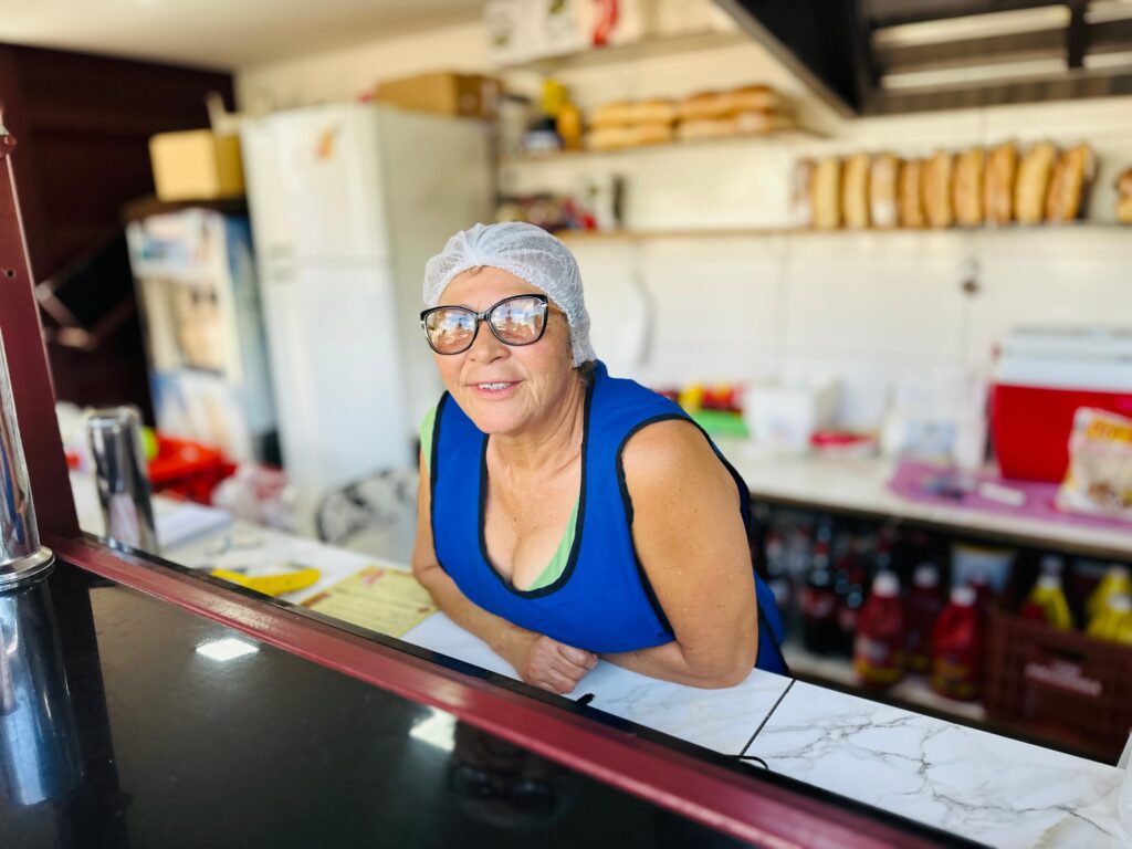 A mulher está vestida com um avental azul sobre uma blusa verde, usando uma touca de cabelo. Ela usa óculos pretos grandes com armação e está apoiada em um balcão. Ao fundo, há prateleiras com pães, garrafas e uma geladeira. A iluminação sugere que a foto foi tirada durante o dia.
