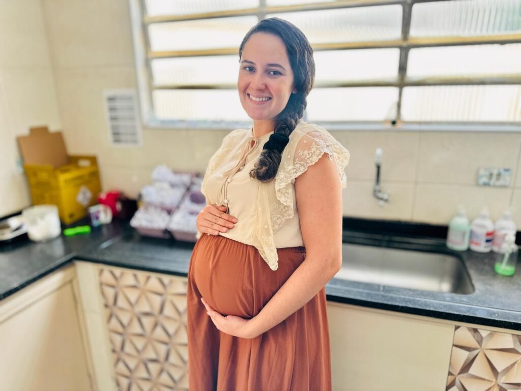 A imagem mostra uma mulher grávida sorrindo para a câmera, com as mãos apoiadas em sua barriga. Ela está vestindo uma blusa bege com detalhes de renda nas mangas e uma saia longa marrom. O fundo sugere que ela está em uma cozinha ou área de serviço, com uma pia, armários e alguns objetos em uma bancada. Há uma caixa amarela com símbolos de perigo à esquerda e recipientes com itens brancos empilhados ao fundo. As janelas acima da pia têm venezianas, e a bancada tem um padrão geométrico decorativo. A iluminação é suave, vindo de cima e da frente, destacando a mulher.