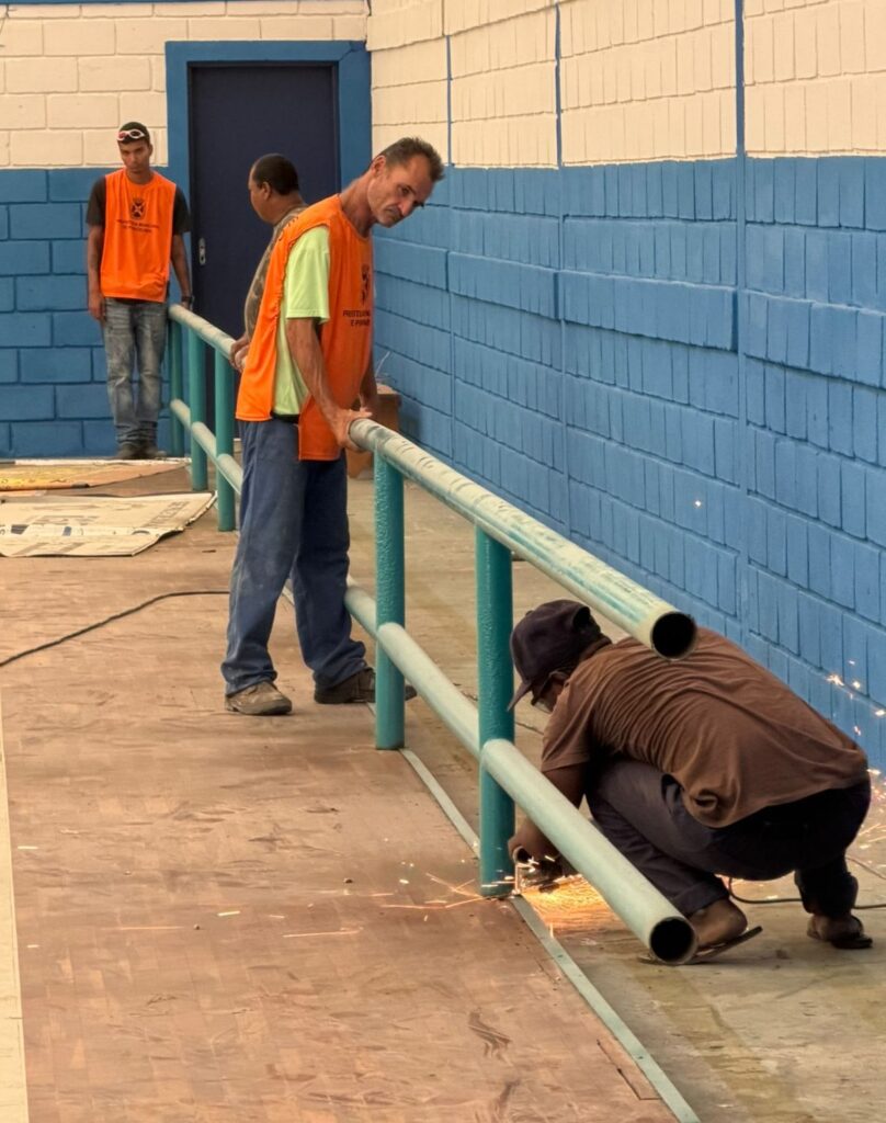 A foto mostra quatro trabalhadores atuando na instalação ou reparo de um corrimão metálico em um ambiente interno com paredes de tijolos pintadas em azul e branco. Um deles utiliza uma ferramenta elétrica que solta faíscas, enquanto dois colegas seguram e estabilizam a estrutura, e outro observa ao fundo. Todos vestem coletes de segurança laranja, e o chão está protegido por lonas e painéis de madeira. A cena transmite a ideia de trabalho em equipe e esforço manual em um contexto de manutenção ou construção.