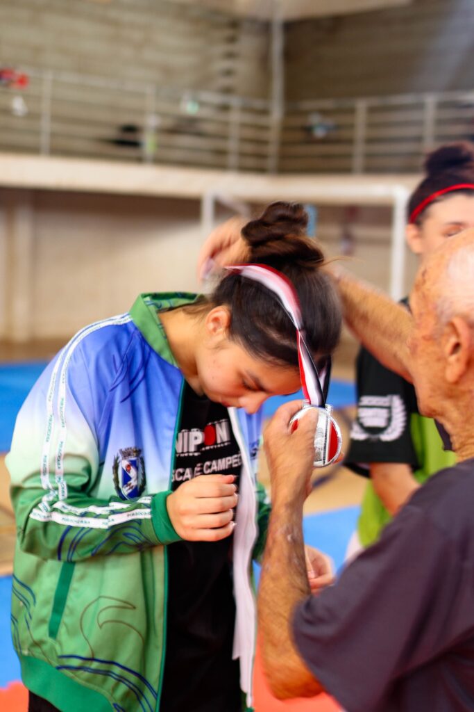 A imagem mostra um momento de premiação em um evento esportivo. Uma jovem, com o cabelo preso em um coque e usando uma faixa vermelha na cabeça, está recebendo uma medalha. Ela veste uma jaqueta esportiva nas cores verde e azul, com detalhes em branco. Um homem mais velho, com a pele enrugada e braços musculosos, está entregando a medalha para ela. Ao fundo, outra jovem com cabelo preso em coque e faixa vermelha na cabeça observa a cena. O ambiente parece ser uma quadra esportiva, com piso azul e grades ao fundo. A atmosfera é de celebração e reconhecimento.