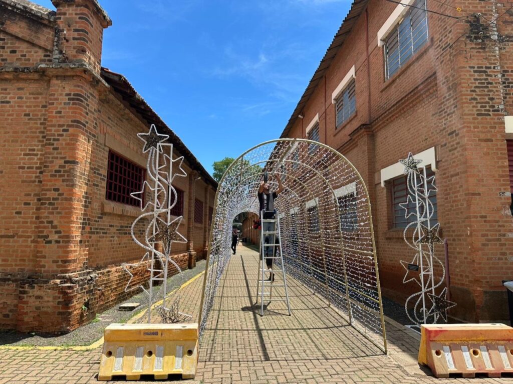 A imagem mostra um túnel decorado com luzes brancas, que parece estar sendo montado ou desmontado. Uma pessoa está em cima de uma escada, trabalhando na estrutura do túnel. Ao lado do túnel, há decorações em forma de estrela com espirais. O cenário é composto por edifícios de tijolos vermelhos com janelas gradeadas e um céu azul claro com algumas nuvens. Há também barreiras de segurança amarelas no chão.