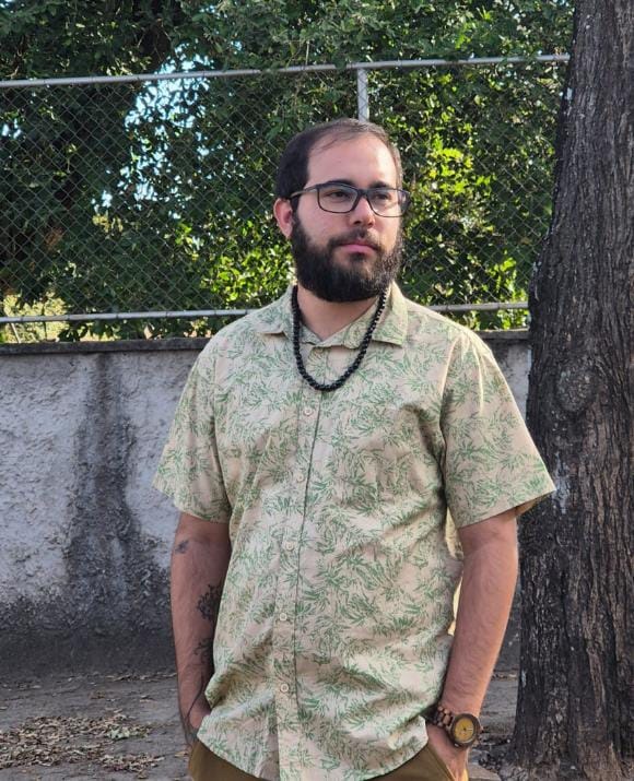 A imagem mostra um homem em pé, com as mãos nos bolsos, vestindo uma camisa de manga curta com estampa de folhas verdes sobre um fundo bege. Ele usa óculos de aro escuro, tem barba e usa um colar de contas pretas. No pulso direito, ele usa um relógio com pulseira de madeira e mostrador redondo. O homem está posicionado em frente a uma parede de concreto e uma cerca de metal, com árvores e folhagens ao fundo. A iluminação sugere que a foto foi tirada durante o dia, possivelmente ao ar livre. Há um tronco de árvore grande à direita do homem, parcialmente visível na imagem.