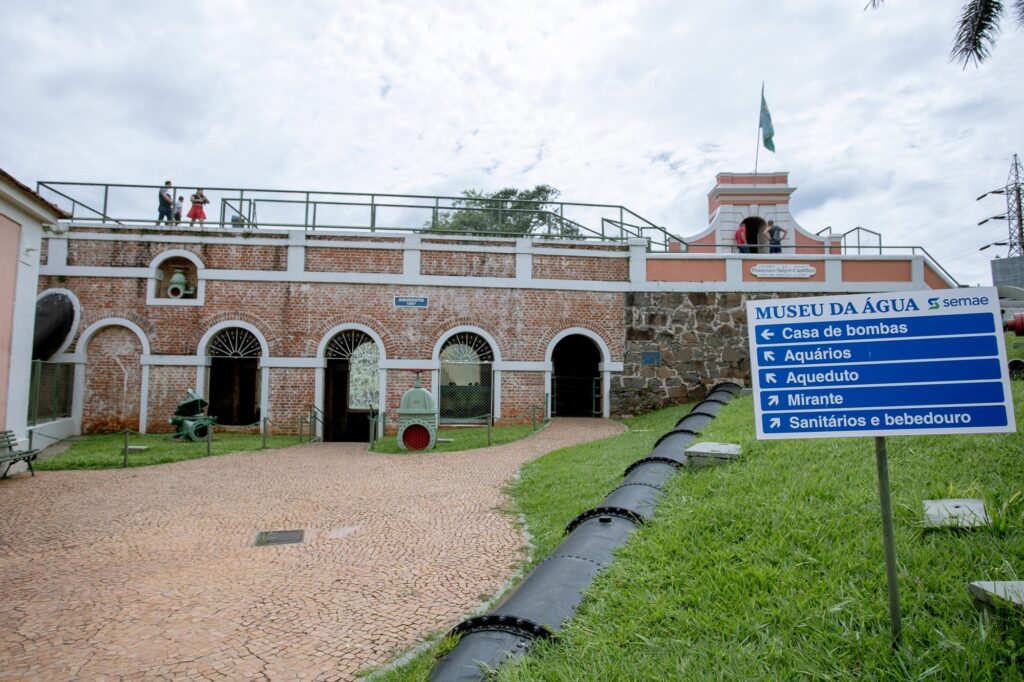 A imagem mostra o exterior de um museu, provavelmente dedicado à água, como indicado pela placa que diz "MUSEU DA ÁGUA". O edifício em si é feito de tijolos com entradas e janelas em arco. Há várias peças grandes e antigas de válvulas de água verdes ou maquinário expostas no terreno. Um grande cano preto corre ao longo da encosta gramada em primeiro plano. À direita do edifício, uma placa azul com texto branco fornece indicações para várias atrações dentro do complexo do museu. Ela lista "Casa de bombas", "Aquários", "Aqueduto", "Mirante" e "Sanitários e bebedouro". No topo da estrutura de tijolos, há uma passarela com corrimão, e algumas pessoas podem ser vistas caminhando por lá. Uma pequena estrutura semelhante a uma torre com uma bandeira é visível no lado direito do nível superior do edifício. O céu está nublado.