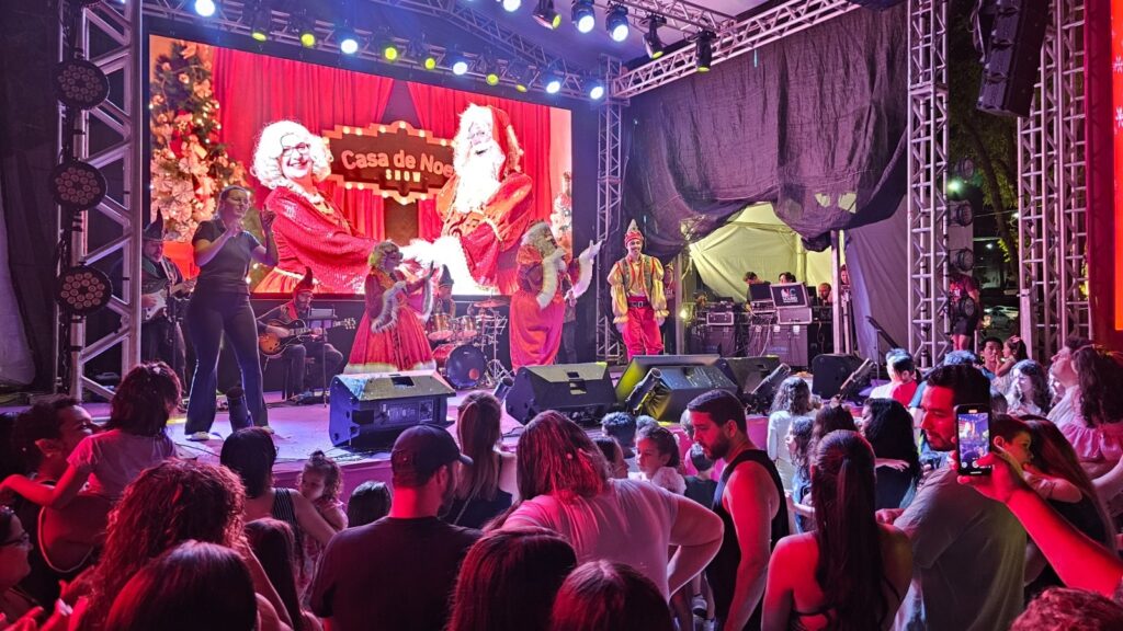 A imagem mostra uma cena vibrante de um show ao vivo em um palco, com uma grande tela de LED ao fundo exibindo uma imagem natalina com a logo "Casa de Noel Show". No palco, há artistas fantasiados, incluindo o Papai Noel e outros personagens festivos, além de músicos tocando instrumentos como guitarra e bateria. Uma mulher em primeiro plano, vestida com roupas casuais, parece estar interagindo com o público ou apresentando o show. A plateia, composta por pessoas de todas as idades, está em frente ao palco, algumas em pé e outras sentadas, aproveitando o espetáculo. A iluminação é colorida e intensa, com predominância de tons vermelhos e roxos, criando uma atmosfera animada e festiva. Há também estruturas metálicas de suporte para o palco e equipamentos de som, indicando que se trata de um evento profissional. A presença de árvores e decorações natalinas sugere que o show está relacionado às celebrações de fim de ano.