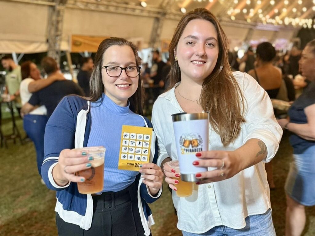 A imagem mostra duas mulheres sorrindo para a câmera em um evento ao ar livre, possivelmente um festival de cerveja, dado o copo e o copo térmico que elas estão segurando. A mulher à esquerda usa óculos, um suéter azul claro e um casaco azul com detalhes brancos. Ela segura um copo de plástico com uma bebida âmbar e um folheto amarelo com ícones. A mulher à direita está usando uma camisa branca e jeans, e segura um copo térmico branco com um logotipo que parece ser de cerveja. O fundo está um pouco desfocado, mas mostra outras pessoas e tendas, sugerindo uma multidão e uma atmosfera festiva.