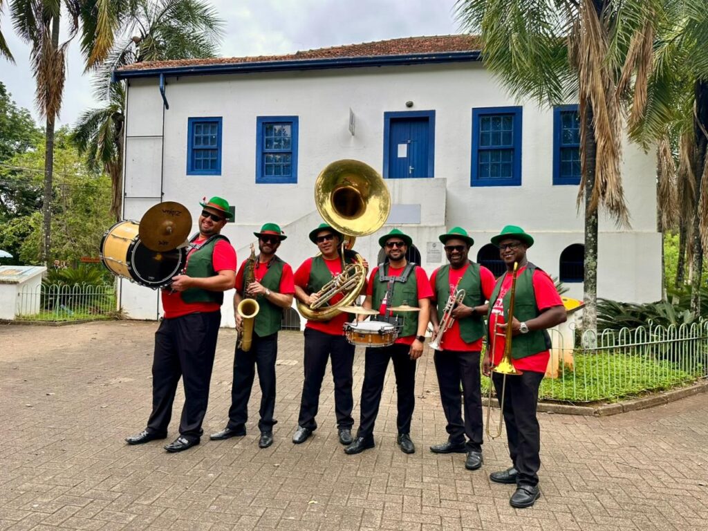 A imagem mostra um grupo de sete homens vestidos com chapéus verdes pontudos, camisetas vermelhas e coletes verdes, posando em frente a um edifício branco com janelas azuis. Eles parecem ser uma banda de metais, pois vários homens seguram instrumentos musicais, incluindo um bumbo, um saxofone, um trombone e um tuba. A banda está em uma área pavimentada com pedras, com árvores e folhagens ao fundo. O céu está nublado, sugerindo um dia fresco.