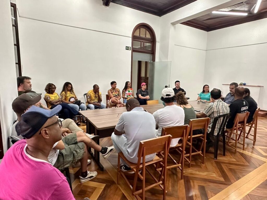 A imagem mostra um grupo de pessoas reunidas em torno de uma mesa de madeira em uma sala com paredes brancas e piso de madeira com um padrão geométrico. A maioria das pessoas está sentada, voltada para a mesa, e algumas parecem estar conversando ou ouvindo. No lado esquerdo da imagem, um homem de camisa rosa e boné azul está em primeiro plano, olhando para a direita. Atrás dele, há outras pessoas sentadas, algumas vestindo camisetas amarelas com estampas. No lado direito da mesa, há um grupo de pessoas sentadas, incluindo um homem com uma camiseta preta com a palavra "COACH" escrita. Uma mulher está sentada em frente a ele, vestindo uma blusa azul clara. A sala parece ter uma arquitetura clássica, com um teto de madeira escura e uma janela arqueada ao fundo. Há também um ventilador de teto visível. A iluminação é suave, vinda de cima. A atmosfera geral sugere uma reunião, palestra ou discussão em grupo.
