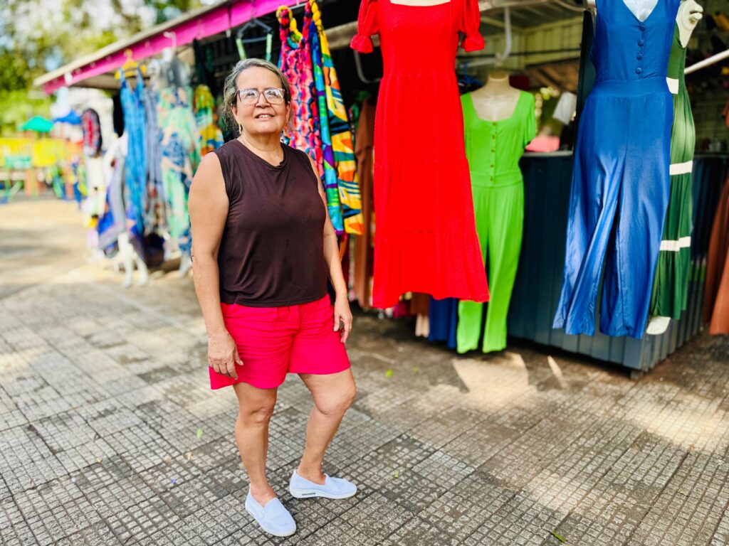 A imagem mostra uma mulher sorrindo para a câmera em frente a uma barraca de roupas coloridas. Ela está usando um top marrom sem mangas e shorts rosa vibrante, combinados com tênis brancos. Ao fundo, há uma variedade de roupas penduradas em cabides, incluindo vestidos e macacões em cores vivas como vermelho, verde e azul. O chão é pavimentado com um padrão quadriculado de pedras. A iluminação sugere um dia ensolarado, com algumas sombras projetadas no chão.