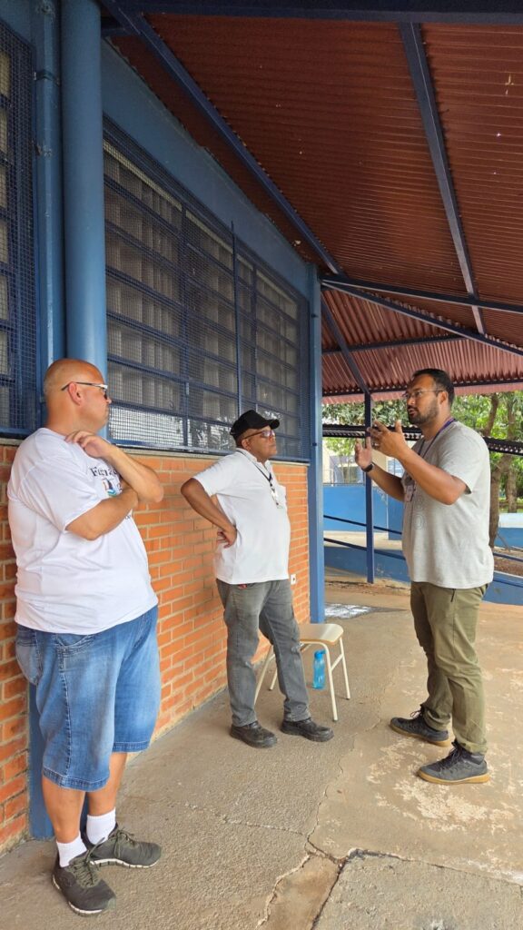 A imagem mostra três homens conversando em pé do lado de fora de um prédio. O homem mais à esquerda é careca, usa óculos escuros, uma camiseta branca e shorts jeans. Ele está encostado em uma parede de tijolos vermelhos. O homem do meio usa um boné preto, óculos, uma camiseta branca e calças jeans cinzas. Ele tem uma mão apoiada na cintura. O homem mais à direita usa óculos, uma camiseta cinza e calças verdes. Ele está gesticulando com as mãos enquanto fala com o homem do meio. Há uma cadeira branca e uma garrafa de água azul no chão atrás dos dois homens mais à direita. O prédio atrás deles tem janelas grades e um telhado de metal cor de ferrugem. O chão é de concreto rachado.