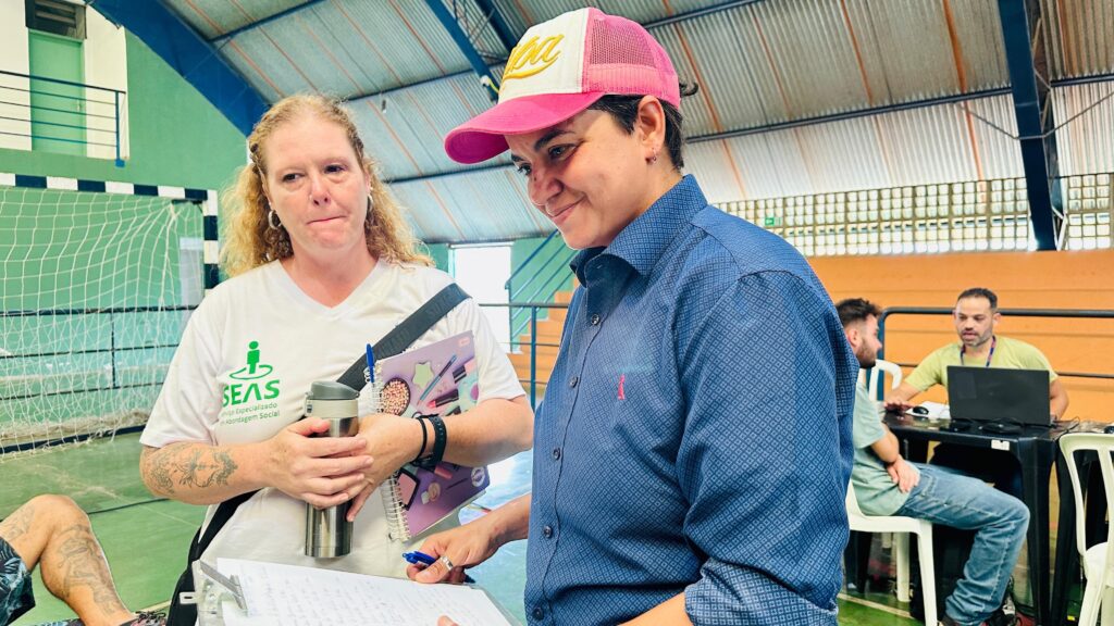 A imagem mostra duas mulheres em primeiro plano, dentro de um ginásio. A mulher à esquerda tem cabelos loiros ondulados e veste uma camiseta branca com um logotipo verde que diz "SEAS Serviço Especializado em Abordagem Social". Ela segura uma caneca térmica prateada e um caderno. Tatuagens são visíveis em seu braço. A mulher à direita usa um boné rosa e branco e uma camisa azul com um padrão xadrez pequeno e um pequeno logo rosa. Ela está sorrindo e olhando para baixo, segurando uma caneta sobre um papel. Ao fundo, em uma mesa, dois homens estão sentados. Um deles, com uma camiseta verde clara, está olhando para um laptop. O outro homem, com uma camiseta azul clara, está conversando com ele. O ambiente parece ser uma quadra esportiva, com uma rede de vôlei visível ao fundo.