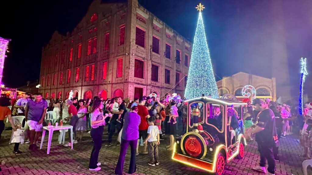 A imagem retrata um evento noturno em frente a um edifício histórico, com uma atmosfera festiva de Natal. * Um imponente pinheiro de Natal, adornado com milhares de luzes azuis, domina o centro da cena, coroado por uma estrela brilhante. * Ao lado da árvore, um pequeno trem decorado com luzes neon, pronto para levar crianças em um passeio encantado. * Uma multidão de pessoas, incluindo famílias com crianças, se reúne, aproveitando o ambiente. * O edifício histórico ao fundo, com suas janelas iluminadas em tons vibrantes de vermelho, adiciona um toque dramático e festivo à paisagem. * A iluminação geral, com luzes coloridas e neon, cria um espetáculo visual cativante, ideal para celebrações de fim de ano.