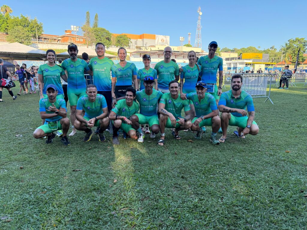 A imagem mostra um grupo de pessoas, vestindo uniformes de ciclismo nas cores verde e azul com o logo "care club" e a frase "your best move". Eles estão em uma área gramada, possivelmente após uma competição ou evento esportivo, com uma estrutura de arquibancadas e antenas ao fundo. A maioria das pessoas está agachada para uma foto em grupo, sorrindo para a câmera. Alguns homens e mulheres do grupo estão em pé atrás dos agachados. A iluminação sugere que a foto foi tirada durante o dia, com o sol iluminando a cena. Há também algumas cercas e equipamentos esportivos visíveis no fundo, indicando um local de evento.