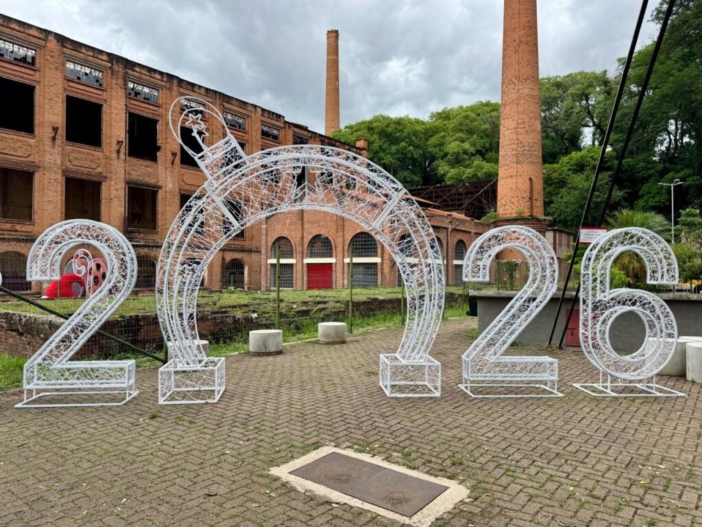 A imagem exibe uma instalação decorativa em frente a um edifício histórico de tijolos, com chaminés altas ao fundo. A instalação consiste em grandes números brancos luminosos, formando "2026", com um arco decorado com uma estrela entre os números "0" e "2". O edifício, com janelas quebradas e aspecto envelhecido, contrasta com a modernidade da iluminação festiva. O chão é pavimentado com tijolos e há vegetação e postes de luz ao redor. O céu está nublado, sugerindo um dia chuvoso ou nublado.