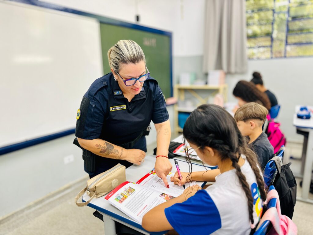 A imagem mostra uma sala de aula onde uma policial fardada, com óculos coloridos e cabelo loiro preso em um rabo de cavalo, está auxiliando uma aluna com seu trabalho. A policial aponta para um livro aberto na mesa da menina, que está escrevendo com uma caneta rosa. Outras crianças estão sentadas em suas carteiras ao fundo, também focadas em suas atividades. A sala de aula parece ser um ambiente escolar típico, com quadros brancos, carteiras e prateleiras com materiais.