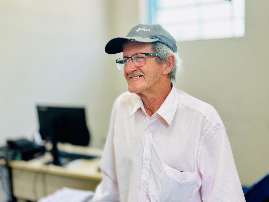 Um homem mais velho, com cabelos grisalhos e bigode, usa um boné cinza com a palavra "sphere" escrita em branco e óculos de aro preto. Ele está vestindo uma camisa social rosa clara desabotoada no colarinho. Ele está sorrindo e olhando para a direita. Ao fundo, um monitor de computador preto em uma mesa, com alguns papéis espalhados. O ambiente parece ser um escritório.