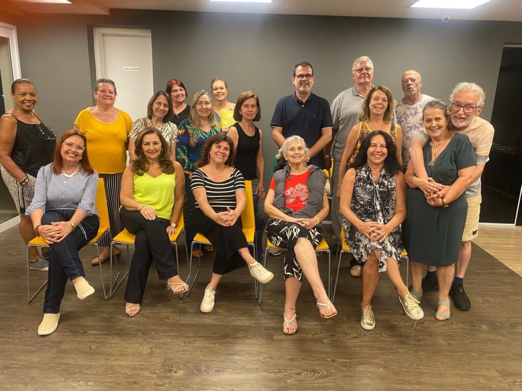 A imagem mostra um grupo de 16 pessoas, divididas em duas fileiras, posando para uma foto em um ambiente interno. A maioria das pessoas é adulta e parece estar em um evento social ou reunião. Na primeira fileira, da esquerda para a direita, vemos uma mulher com um top preto e saia estampada, sentada em uma cadeira amarela. Ao lado dela, uma mulher com um top azul claro e calça jeans escura, também sentada. Em seguida, uma mulher com um top amarelo vibrante e calça preta, sentada. Mais à direita, uma mulher com um top verde limão e calça preta, sentada. Uma mulher com um top estampado em cores vibrantes, sentada. Outra mulher com um top preto e branco listrado, sentada. E por último, uma mulher com um vestido verde escuro, sentada. Na segunda fileira, em pé, da esquerda para a direita, vemos uma mulher com um top preto e saia estampada. Ao lado dela, uma mulher com um top amarelo. Uma mulher com um top estampado em tons de azul e verde. Uma mulher com um top preto. Mais ao centro, um homem com camisa azul escura. Ao lado dele, um homem com camisa cinza. Uma mulher sorridente com um top amarelo. Um homem com uma camisa estampada. E à direita, um homem com camisa bege e óculos, sorrindo. O fundo da imagem é uma parede cinza, com uma porta branca à esquerda e uma luminária no teto. O chão é de madeira clara. A iluminação é suave, vindo de cima.