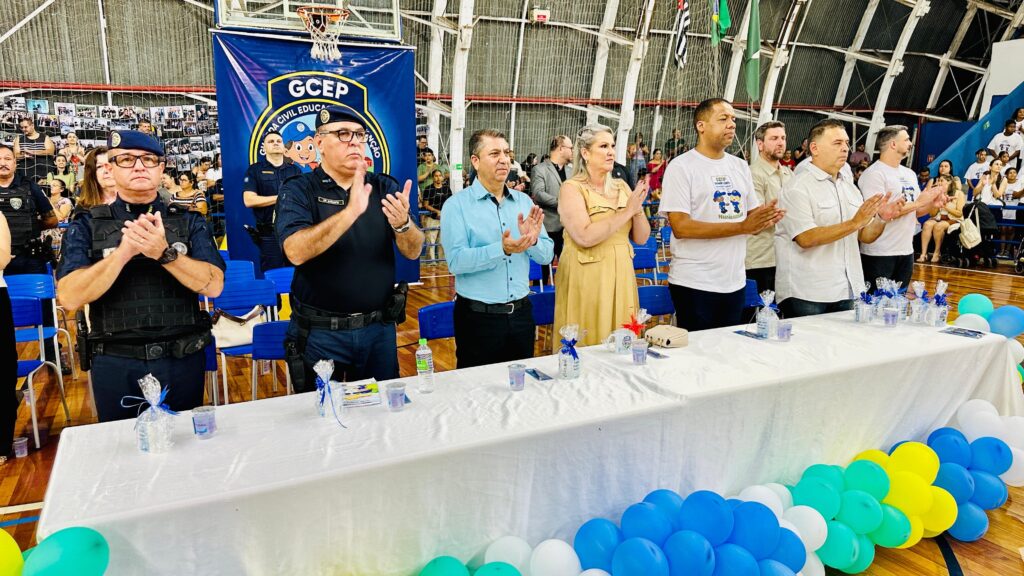 A imagem mostra um evento em um ginásio, possivelmente uma formatura ou cerimônia. Há várias pessoas em pé atrás de uma mesa coberta com uma toalha branca. No centro da mesa, há pequenos pacotes embrulhados em celofane azul e branco, alguns copos e uma garrafa de água. Na frente da mesa, há uma fileira de balões coloridos, predominantemente azuis, brancos e amarelos, formando um arco. As pessoas atrás da mesa parecem estar aplaudindo. Várias bandeiras estão penduradas ao fundo, incluindo a bandeira do Brasil e a bandeira de São Paulo. Há também um grande banner azul com o logotipo "GCEP" e desenhos animados. À esquerda, dois homens uniformizados, aparentemente policiais, estão de pé e aplaudindo. Um deles usa um colete tático. À direita, outras pessoas em trajes civis também aplaudem. Ao fundo, pode-se ver a arquibancada com espectadores.