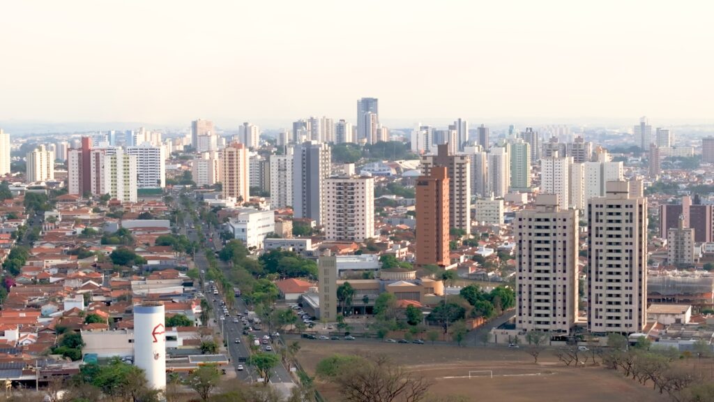 A imagem mostra uma vista aérea de uma grande cidade durante o dia, com uma luz suave que sugere início da manhã ou fim da tarde. A cidade é bastante densa, com muitos prédios altos modernos, além de casas mais antigas com telhados de cerâmica vermelha. No centro da imagem, uma avenida principal arborizada corta a cidade, com carros circulando. Em primeiro plano, destaca-se um grande reservatório de água branco com um logo vermelho. A paisagem urbana se estende até o horizonte, onde o contorno dos prédios começa a se perder na névoa, transmitindo a ideia de uma cidade muito grande. A combinação de diferentes estilos arquitetônicos e a presença de áreas verdes dão um aspecto vibrante e diversificado ao cenário. A imagem transmite uma sensação de movimento e vida urbana, apesar de não mostrar pessoas diretamente.