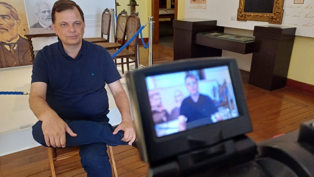 A imagem mostra um homem sentado em uma cadeira de madeira, vestindo uma camisa polo azul escura e calças jeans. Ele está em um ambiente que parece ser um museu ou galeria, com quadros e expositores ao fundo. Há também um tripé de câmera com uma tela visível, exibindo uma imagem desfocada de alguém. O homem olha para a câmera com uma expressão neutra. A iluminação é suave, destacando o homem e o ambiente ao seu redor.
