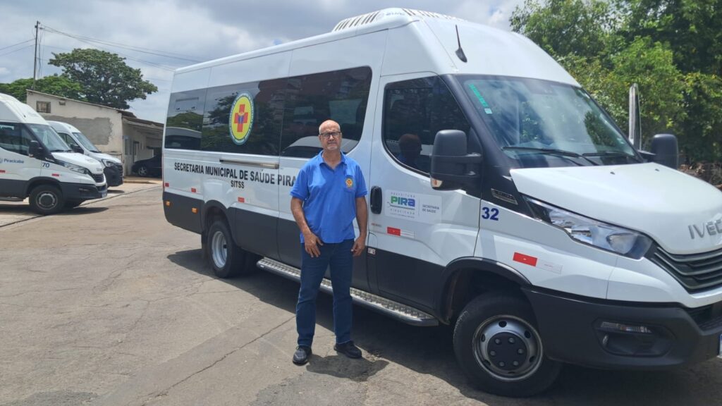 A imagem mostra um homem de óculos e camisa azul em pé ao lado de uma van branca. A van tem a inscrição "SECRETARIA MUNICIPAL DE SAÚDE PIRA" e o número "32" pintado na lateral. Há também um logotipo com uma cruz vermelha dentro de um círculo amarelo. Ao fundo, outras vans brancas semelhantes estão estacionadas, e árvores verdes e um céu azul podem ser vistos. O homem parece estar em um estacionamento ou pátio, com o asfalto rachado no chão.