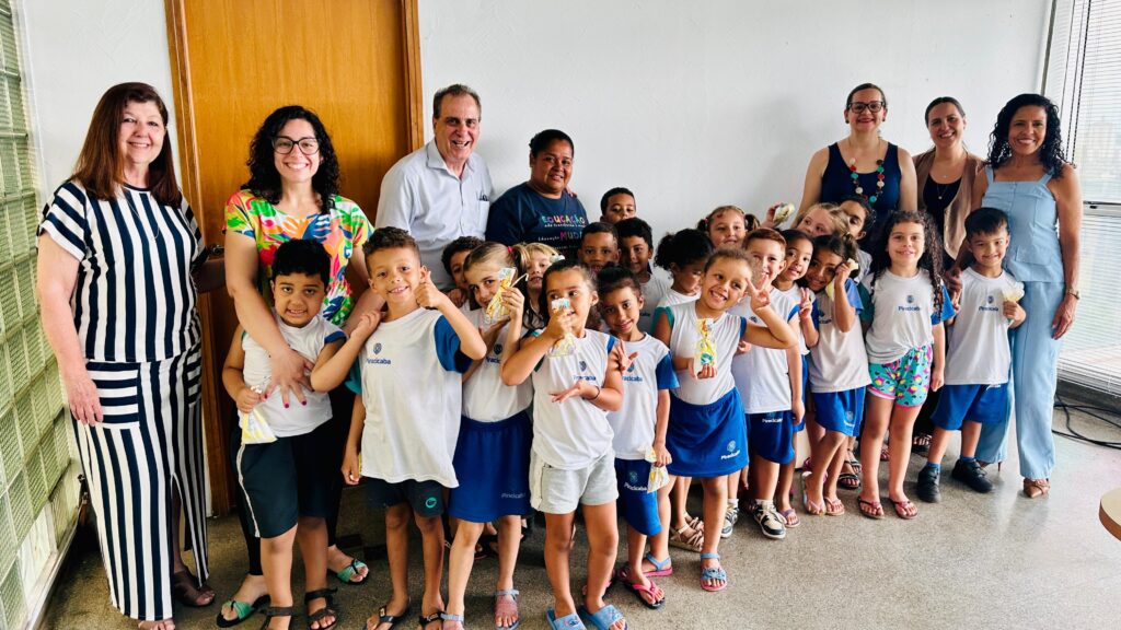 A imagem mostra um grupo de adultos e crianças em um ambiente interno, possivelmente uma escola. A maioria das crianças está vestida com uniformes escolares brancos com detalhes azuis e seguram pequenos pacotes embrulhados. Os adultos, que parecem ser professores ou funcionários da escola, estão atrás ou ao lado das crianças, sorrindo para a câmera. No lado esquerdo da imagem, uma mulher mais velha com cabelo castanho escuro e uma mulher mais jovem com cabelo castanho claro e óculos estão em pé. Atrás delas, um homem de camisa branca sorri. No centro, há um grupo de crianças, algumas segurando pacotes, e atrás delas, uma mulher com uma camiseta azul escuro com a inscrição "EDUCAÇÃO NÃO TRANSFORMA MUDANDO". No lado direito, há mais adultos, incluindo duas mulheres com óculos e uma mulher com cabelo escuro e um vestido azul claro.