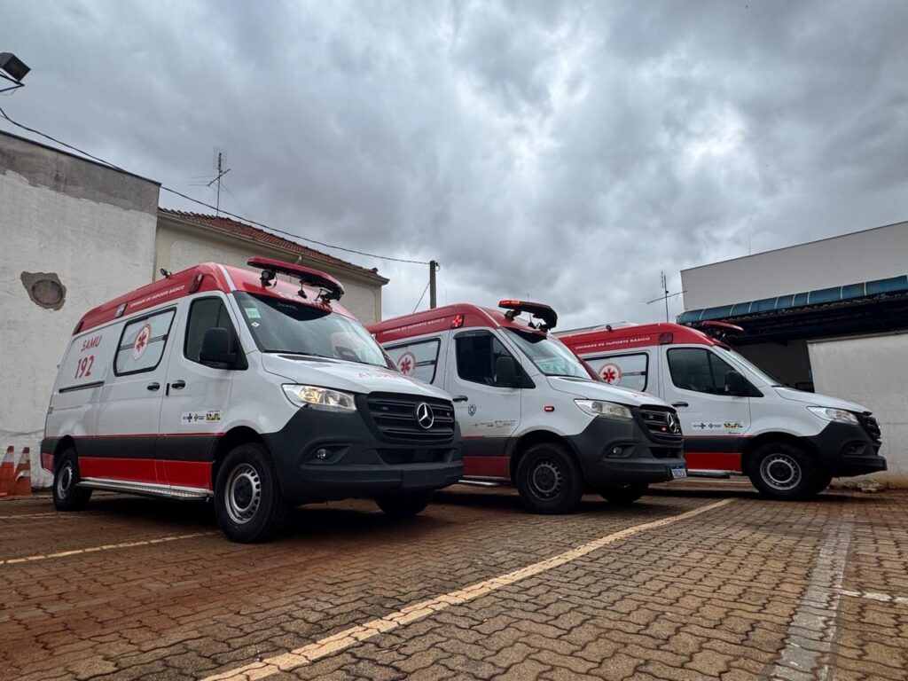 A imagem mostra três ambulâncias brancas com detalhes vermelhos estacionadas em uma área pavimentada. As ambulâncias estão alinhadas, sendo a da esquerda a mais próxima e em foco. Todas têm "SAMU 192" escrito na lateral, que é o Serviço de Atendimento Móvel de Urgência do Brasil. As ambulâncias também possuem uma luz vermelha no topo e uma faixa vermelha na parte inferior. O fundo consiste em prédios e um céu nublado. A impressão geral é de prontidão e serviços de emergência.