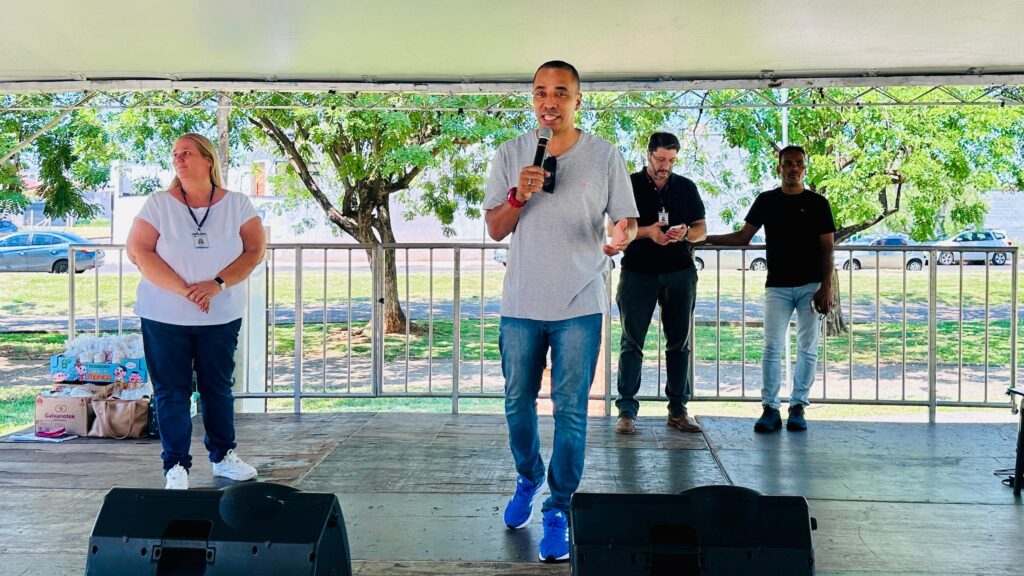 A imagem mostra um palco ao ar livre com três pessoas em pé. No centro, um homem de pele morena, vestindo uma camiseta cinza e calça jeans, segura um microfone e parece estar falando. À sua esquerda, uma mulher loira, com uma camiseta branca e calça jeans escura, está de pé com as mãos cruzadas. À direita do orador principal, há dois homens. Um deles, vestindo uma camiseta preta e calça jeans, está ligeiramente atrás e parece estar olhando para um celular. O outro homem, também de camiseta preta e calça jeans clara, está mais à direita e parece estar observando a plateia. Atrás deles, vê-se uma área com árvores e carros estacionados. Na frente do palco, há caixas e sacolas, possivelmente com doações ou materiais para um evento.