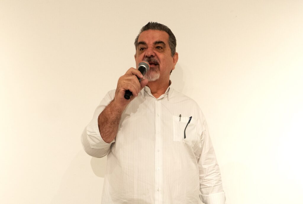 A imagem mostra um homem de meia-idade, com cabelos escuros e barba grisalha, vestindo uma camisa branca com listras finas. Ele está segurando um microfone preto com a mão direita e falando. A camisa tem um bolso no lado esquerdo do peito, onde há uma caneta preta e um par de óculos escuros com armação preta. O fundo da imagem é uma parede branca lisa. A iluminação é suave, destacando o rosto do homem e a textura da camisa.