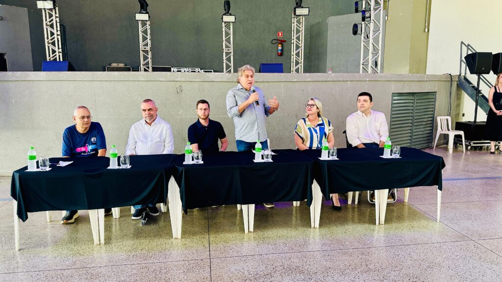 A imagem mostra um grupo de seis pessoas sentadas atrás de uma longa mesa coberta com uma toalha preta. Há garrafas de água e copos na mesa. À esquerda, um homem com óculos e uma camiseta azul escura com um laço rosa está sentado. Ao lado dele, um homem com uma camisa branca e barba está sentado. No centro, um homem de camisa cinza e jeans está em pé, segurando um microfone e falando. Ao lado dele, uma mulher com óculos e uma blusa colorida está sentada. À direita, um homem de camisa branca está sentado. Mais à direita, uma mulher com um vestido preto está em pé. Ao fundo, há uma estrutura de palco com luzes e equipamentos de som. Uma escada leva a uma plataforma mais alta. Há também uma cadeira branca e uma porta de metal no fundo. O chão é cimentado.
