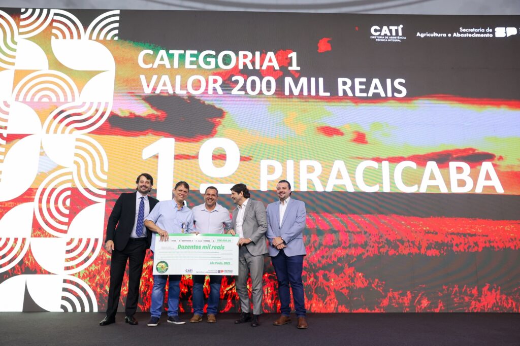 A imagem mostra um evento de premiação, onde cinco homens estão em pé em frente a um grande telão. O telão exibe um fundo vibrante com um padrão geométrico abstrato no lado esquerdo e uma paisagem rural em tons de vermelho e laranja no lado direito. Texto em letras grandes e brancas domina a tela, indicando "CATEGORIA 1", "VALOR 200 MIL REAIS", "1º" e "PIRACICABA". No centro da imagem, quatro dos homens estão reunidos em torno de um cheque gigante. Dois deles, vestindo camisas azuis, seguram o cheque. Um homem de terno preto está à esquerda, e outro de terno cinza está à direita dos que seguram o cheque. O quinto homem, vestindo um terno azul claro, está posicionado mais à direita, ligeiramente separado do grupo. O cheque, com um selo verde no canto inferior esquerdo, tem o valor "200.000,00" escrito em numérico e "Duzentos mil reais" por extenso. Abaixo do valor, lê-se "São Paulo, 2025". A logo da CATI (Diretoria de Assistência Técnica Integral) e da Secretaria de Agricultura e Abastecimento do Estado de São Paulo também estão visíveis no telão e no cheque. A atmosfera geral sugere uma celebração de conquista ou reconhecimento financeiro no setor agrícola.