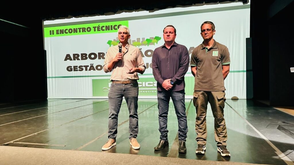 A imagem mostra três homens em pé em um palco, com um banner ao fundo. O homem da esquerda está falando em um microfone e vestindo uma camisa bege, calça jeans e tênis. O homem do meio veste uma camisa social escura e calça jeans escura, com um relógio no pulso. O homem da direita veste uma camisa polo verde, calça cargo e tênis. O banner ao fundo exibe o texto "II ENCONTRO TÉCNICO ARBOR GESTÃO" com ilustrações de árvores e prédios. O palco parece ser feito de madeira escura e há uma iluminação suave vindo de cima.