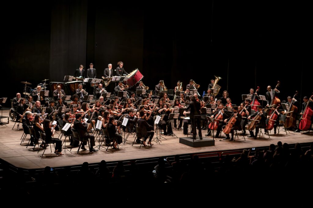 A imagem mostra uma orquestra sinfônica se apresentando em um palco escuro. A iluminação foca nos músicos e em seus instrumentos, criando um contraste dramático com o fundo preto. No palco, músicos de diferentes seções da orquestra estão posicionados. Na frente, há um grupo de violinistas e violoncelistas, seguidos por instrumentos de sopro e metais mais atrás. Um maestro, de costas para o espectador, está em uma plataforma elevada, conduzindo a apresentação com gestos expressivos. Ao fundo, pode-se ver a seção de percussão com tambores e um grande bumbo vermelho. A plateia, embora visível na parte inferior da imagem, está em grande parte escura, com apenas alguns contornos de pessoas e luzes sutis indicando sua presença. A atmosfera geral é de formalidade e concentração, típica de um concerto clássico.