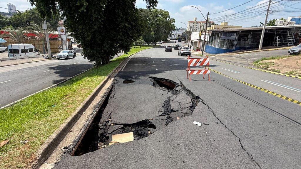 Na imagem, observa-se uma rua da cidade interditada devido a um grande afundamento no asfalto, formando uma cratera profunda. A área danificada está isolada por barreiras do Departamento de Transporte (DOT), sinalizando reparos ou investigação em andamento. O pavimento apresenta rachaduras e colapso evidente, enquanto o tráfego segue normalmente nas faixas adjacentes. Ao redor, veem-se prédios e veículos, compondo o cenário urbano. A cena evidencia um problema de infraestrutura que representa risco à segurança viária e a necessidade urgente de manutenção da via.
