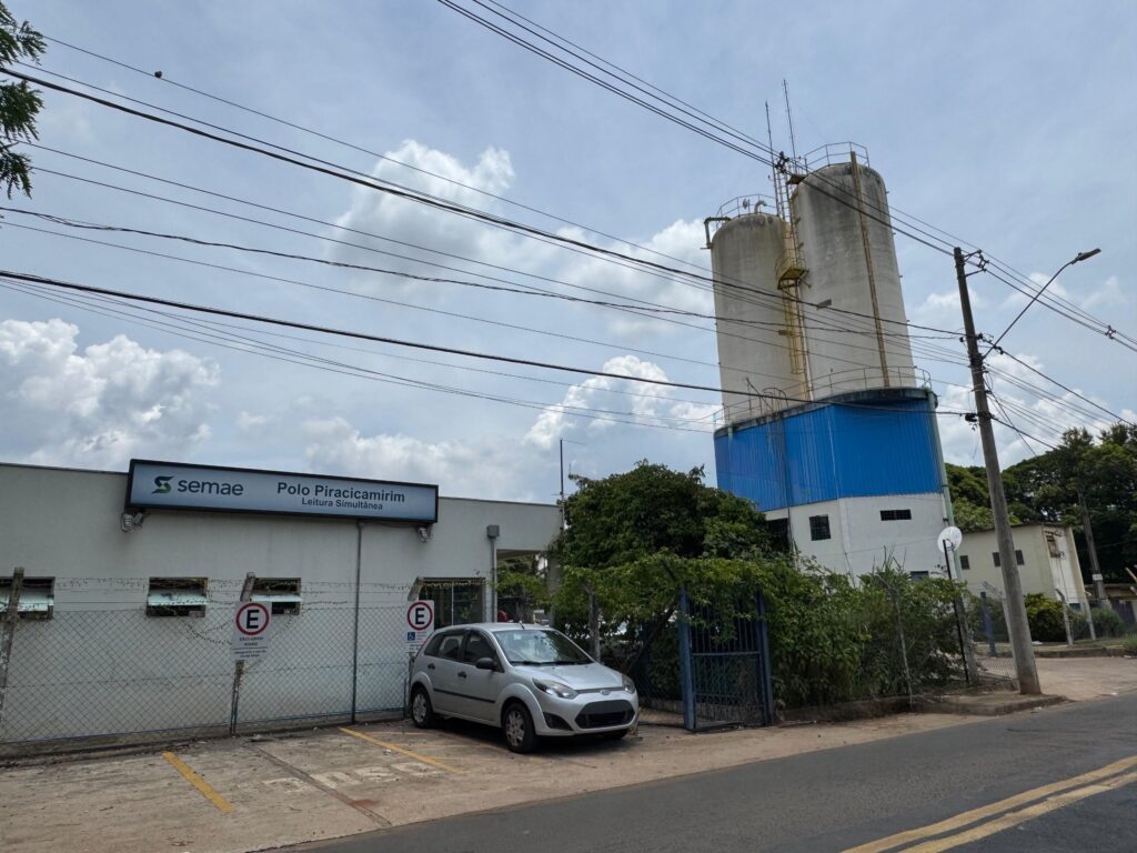 A imagem mostra a fachada de um edifício com uma placa luminosa que diz "semae Polo Piracicamirim Leitura Simultânea". Ao lado do edifício, há um carro prateado estacionado em frente a um portão de metal e uma cerca. Atrás do edifício, erguem-se duas grandes torres cilíndricas de cimento, com uma estrutura azul na base de ambas. O céu está nublado, com nuvens brancas e cinzentas espalhadas. Vários fios elétricos atravessam a parte superior da imagem. Há também postes de iluminação pública e árvores ao redor.