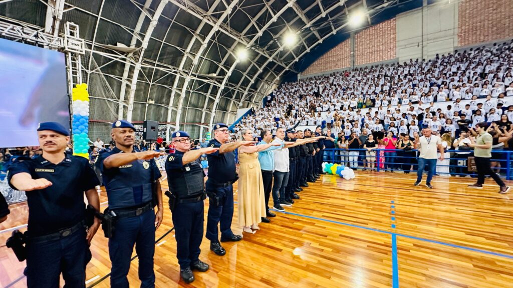 A imagem mostra um evento realizado em uma quadra esportiva com arquibancadas lotadas de pessoas, a maioria vestindo camisetas brancas. Em primeiro plano, um grupo de homens uniformizados, aparentemente policiais, está em posição de sentido, com a mão direita estendida para a frente. Eles usam boinas azuis e uniformes escuros, alguns com coletes táticos. Atrás deles, outros indivíduos em trajes civis também estão em posição de respeito, com as mãos estendidas. O ambiente é de um evento formal, possivelmente uma cerimônia ou formatura, dada a organização das pessoas e a presença de autoridades. A iluminação da quadra é característica de ambientes esportivos, com um teto arqueado e luminárias pendentes. Ao fundo, nas arquibancadas, a multidão observa atentamente o que acontece na quadra. Há também um telão à esquerda, embora seu conteúdo não seja claramente visível. Balões nas cores azul, amarelo e branco estão dispostos em um dos cantos da quadra, adicionando um toque festivo ao evento.