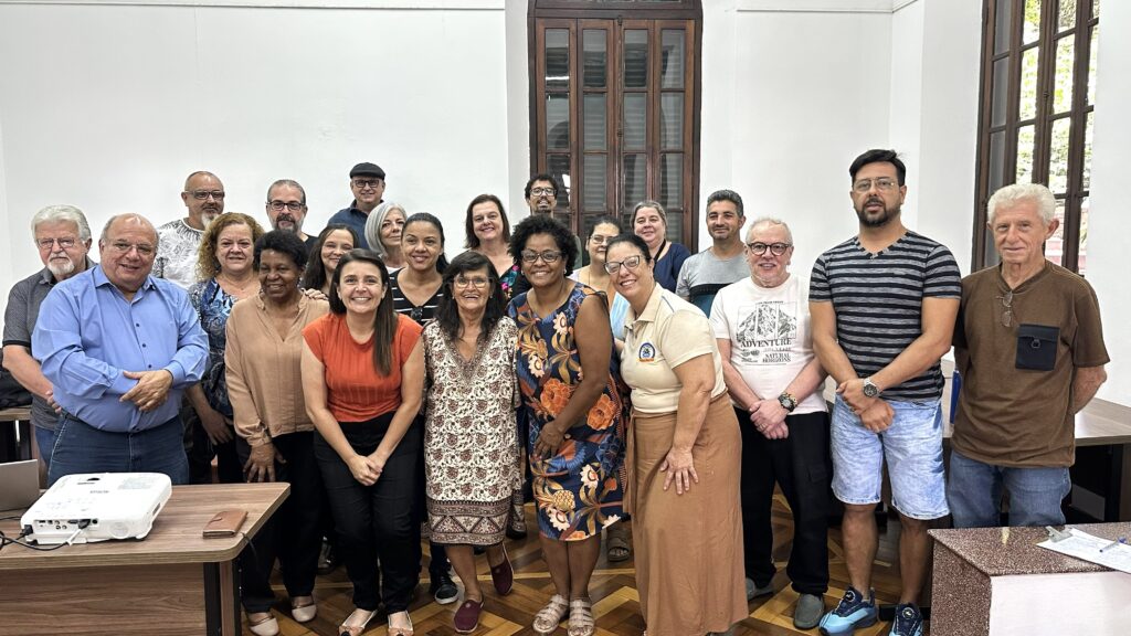 A imagem mostra um grupo de cerca de 20 pessoas, de diversas idades e etnias, posando para uma foto em uma sala. A maioria das pessoas está sorrindo e olhando para a câmera. Há um projetor e uma mesa na frente do grupo, sugerindo que pode ser uma sala de aula ou de reunião. O ambiente parece ser um local fechado, com paredes claras e uma janela grande de madeira escura ao fundo. As roupas das pessoas variam, desde casuais até mais formais, como camisas sociais e vestidos. A iluminação é clara e uniforme, iluminando bem o grupo.