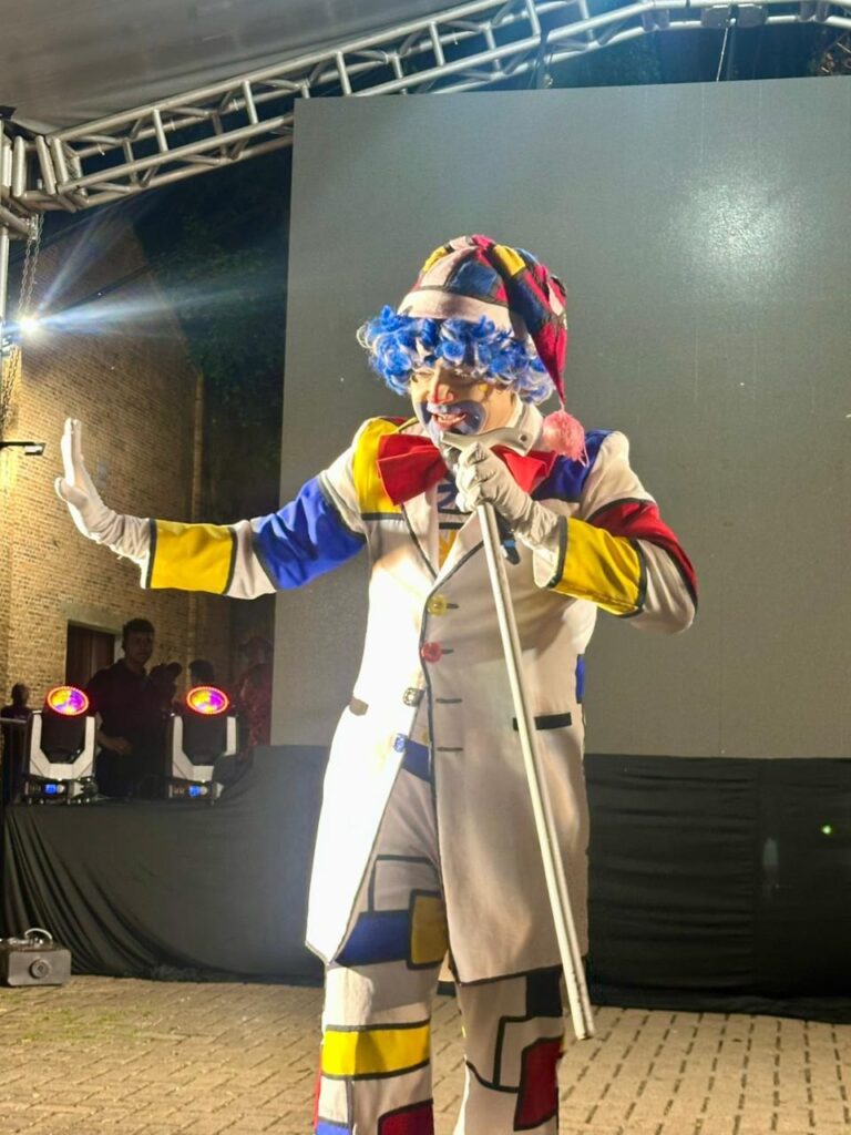 A imagem mostra um palhaço em um palco com uma iluminação dramática. O palhaço está vestindo uma roupa colorida, com um padrão geométrico que lembra as obras de Piet Mondrian, caracterizada por blocos de cores primárias (vermelho, azul e amarelo) e linhas pretas sobre um fundo branco. Ele usa uma peruca azul vibrante e um chapéu listrado em tons de vermelho, azul e amarelo. O rosto do palhaço está pintado com maquiagem tradicional, incluindo um nariz vermelho e detalhes azuis ao redor dos olhos. Ele está segurando um bastão branco em sua mão direita e levantando a mão esquerda em um gesto de saudação ou para chamar a atenção. Ao fundo, pode-se ver uma estrutura metálica de palco com luzes, e uma parede de tijolos à esquerda. À direita, há uma grande tela cinza que pode estar projetando algo ou servindo como pano de fundo. No lado esquerdo inferior da imagem, há equipamentos de som e luz, e algumas pessoas observando. O chão é pavimentado com pedras. A iluminação geral sugere que o evento está ocorrendo à noite.