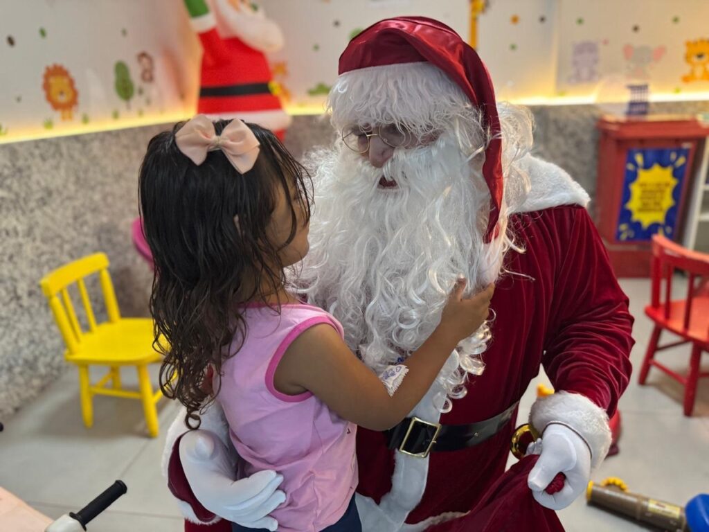 A imagem mostra uma menina abraçando o Papai Noel. A menina, vista de costas, tem cabelos escuros presos com um laço rosa claro e veste uma blusa rosa. Ela está abraçando o Papai Noel, que usa seu traje vermelho tradicional com detalhes brancos e um chapéu. Ele tem uma barba branca longa e óculos. Ao fundo, há cadeiras infantis amarelas e vermelhas, além de decorações temáticas de Natal, como um Papai Noel maior pendurado na parede e desenhos de animais. A cena parece ser um momento afetuoso entre a criança e o personagem natalino.