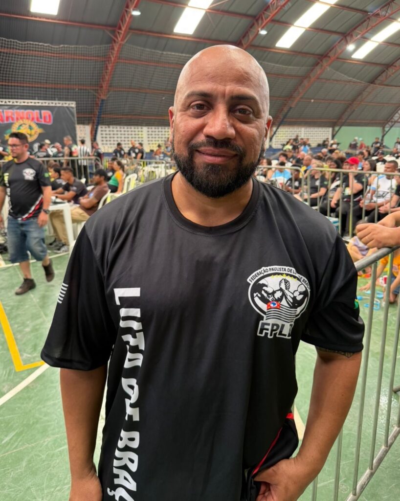 A imagem mostra um homem careca de meia-idade, com barba, vestindo uma camiseta preta com logotipos e texto. Ele está em um evento esportivo, com uma multidão e uma quadra ao fundo. A camiseta tem um logotipo redondo no lado direito do peito com o texto "FEDERAÇÃO PAULISTA DE LUTA DE BRAÇO" e "FPLB" abaixo. No lado esquerdo da camiseta, em letras brancas verticais, está escrito "LUTA DE BRAÇO". O homem parece estar sorrindo levemente para a câmera. O fundo sugere que o evento é uma competição de luta de braço, com pessoas assistindo e competidores em segundo plano. Há uma faixa com o nome "ARNOLD" visível ao fundo, possivelmente indicando o nome do evento ou patrocinador.