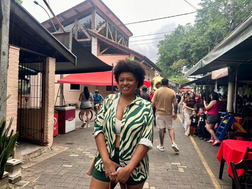 A imagem mostra uma mulher sorridente em primeiro plano, usando uma camisa verde e branca com estampa de zebra e shorts combinando. Ela está em uma rua de paralelepípedos, com edifícios e barracas em ambos os lados. Ao fundo, há outras pessoas passeando, algumas mesas com toalhas vermelhas e uma estrutura de madeira com um telhado inclinado. O céu está nublado, sugerindo um dia com pouca luz solar. A cena parece ser um mercado ou uma área de lazer ao ar livre, com uma atmosfera animada e casual.