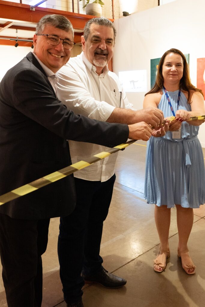A imagem retrata um momento de inauguração ou evento especial com três pessoas em primeiro plano, cortando uma fita. À esquerda, um homem de óculos, terno escuro e camisa clara sorri para a câmera. Ao seu lado, um homem com barba grisalha, camisa branca de botões e um leve sorriso também participa do corte da fita. À direita, uma mulher com um vestido azul claro, alças finas e um crachá preso ao pescoço, segura a fita com as mãos, também sorrindo. A fita a ser cortada é amarela e preta, indicando um evento de abertura. O fundo sugere um ambiente interno, possivelmente uma galeria de arte ou um espaço de eventos, com obras de arte visíveis nas paredes e uma planta em vaso ao fundo. A iluminação é suave, focando nos participantes e no ato do corte da fita.