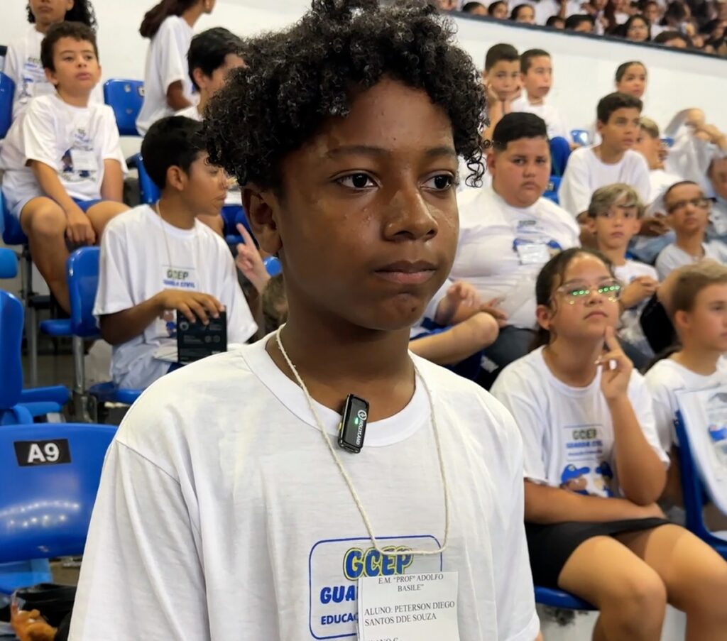 A imagem mostra um grupo de crianças e adolescentes sentados em cadeiras azuis, vestindo camisetas brancas com o logo "GCEP". No centro da imagem, um garoto negro de cabelo cacheado está em primeiro plano, olhando para a direita. Ele usa uma camiseta branca com um crachá pendurado no pescoço, onde se lê "GCEP", "EM. 'PROF ADOLFO BASILE'", "ALUNO: PETERSON DIEGO SANTOS DDE SOUZA" e "EDUCA". Um pequeno microfone preto está preso à sua camiseta. Ao fundo, outros jovens estão sentados, alguns olhando para a frente, outros para os lados. As cadeiras são azuis e algumas têm o número "A9" em um adesivo branco. O ambiente parece ser um auditório ou ginásio.