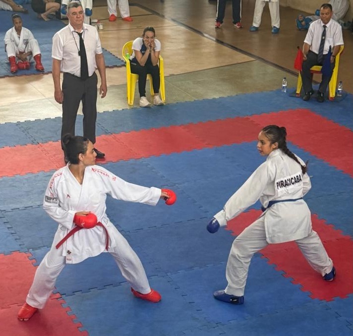 A imagem mostra uma competição de karatê com duas jovens lutadoras em um tatame azul e vermelho. Ambas vestem o tradicional quimono branco, com uma delas usando um cinto vermelho e a outra um cinto azul. Elas estão em posições de combate, com luvas de proteção nas mãos, uma vermelha e outra azul. Ao fundo, um árbitro de camisa branca e calça preta observa atentamente a luta. Sentadas em cadeiras amarelas, duas mulheres assistem à competição, uma delas com expressão concentrada e a outra com as mãos no rosto. Outras pessoas, possivelmente competidores ou espectadores, estão em segundo plano, algumas sentadas e outras em pé, vestindo roupas esportivas. O ambiente sugere uma competição oficial de artes marciais.