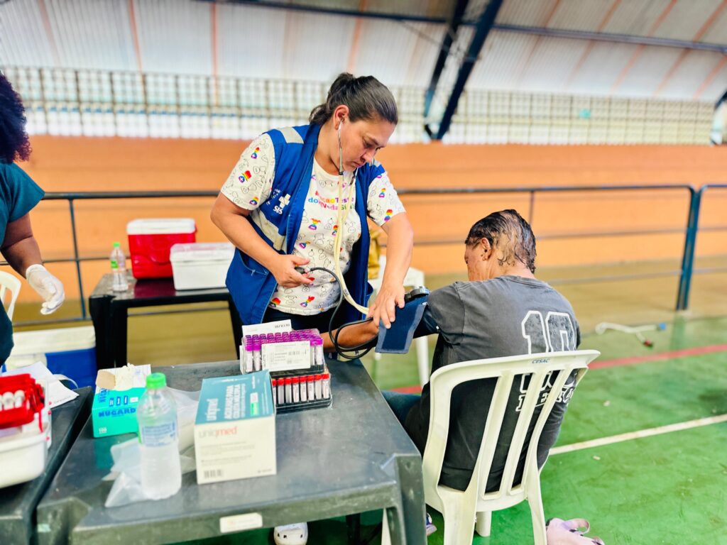 Na imagem, uma profissional de saúde, vestindo um colete azul sobre uma camiseta branca estampada e um estetoscópio ao redor do pescoço, está medindo a pressão arterial de uma pessoa sentada em uma cadeira branca. A profissional segura o manguito do aparelho de pressão no braço do paciente, enquanto observa o medidor. Em uma mesa à frente, há diversos materiais médicos, como tubos de coleta de sangue, caixas de agulhas, luvas e garrafas de água. Ao fundo, observam-se coolers e uma estrutura de arquibancada, sugerindo que o local pode ser uma quadra esportiva ou um local de eventos adaptado para a ação de saúde. Outra pessoa, com luvas, está parcialmente visível à esquerda, indicando que mais de uma pessoa está envolvida na atividade.