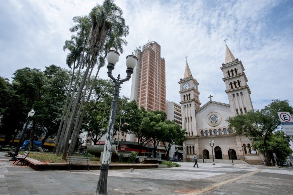 A imagem exibe uma cena urbana com uma igreja de arquitetura clássica ao centro, destacando-se suas torres com relógios e telhados pontiagudos. À esquerda da igreja, ergue-se um edifício moderno de múltiplos andares, com fachada em tons de tijolo e concreto. A composição é complementada por uma praça com palmeiras altas e árvores frondosas, um poste de iluminação com duas lâmpadas e bancos de praça, sugerindo um espaço público convidativo. O céu, com nuvens dispersas, confere um tom mais suave à cena. Um indivíduo caminha pela praça, adicionando um toque de movimento à imagem.