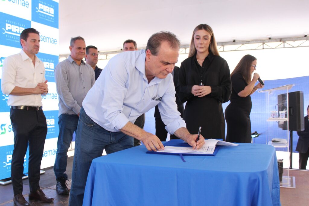A imagem mostra um grupo de pessoas em um evento ao ar livre, possivelmente uma cerimônia de assinatura. No centro da imagem, um homem de camisa azul clara e jeans está se inclinando sobre uma mesa coberta com uma toalha azul, assinando um documento com uma caneta. Ele está focado em sua tarefa. À esquerda do homem que assina, há outro homem vestindo uma camisa branca impecável e calças pretas, com as mãos juntas na frente do corpo. Atrás dele, outros homens estão em pé, observando a cena. À direita do homem que assina, uma mulher de vestido preto está em pé, com as mãos cruzadas na frente do corpo, e ao lado dela, outra mulher de preto está em pé atrás de um púlpito transparente, segurando um microfone e falando. O fundo da imagem é um painel azul com logotipos e texto, incluindo "PREFEITURA" e "PIRACICABA". A iluminação sugere que o evento está ocorrendo durante o dia.